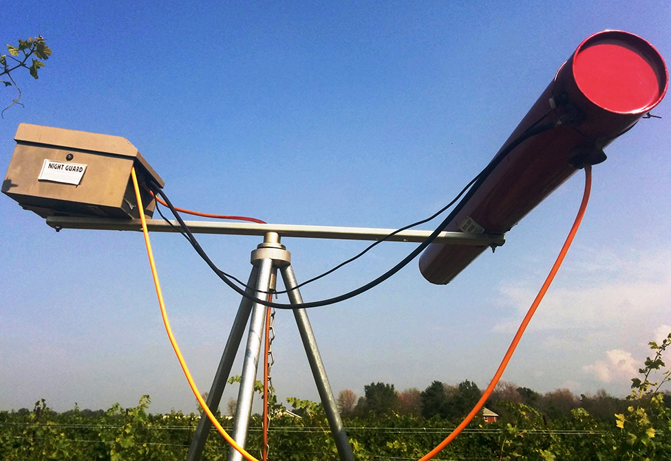 un canon au gaz propane fait d’un trépied avec une barre en travers dans la partie supérieure. Une boîte est fixée sur le côté gauche de la barre et un long tube est fixé sur le côté droit de la barre. Des câbles relient la boîte au tube.