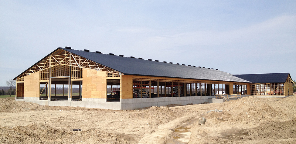 Photo d’une grande grange rectangulaire avec un toit noir en construction. On voit l’ossature des murs et les chevrons.
