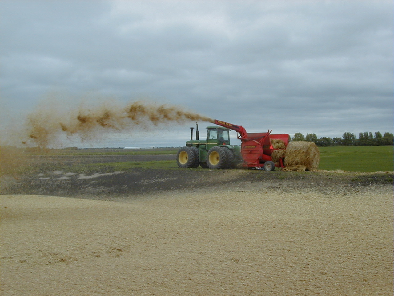 Tracteur auquel est fixé un accessoire qui hache une balle de paille et souffle la paille hachée sur la surface du fumier liquide dans une structure de stockage de fumier en terre