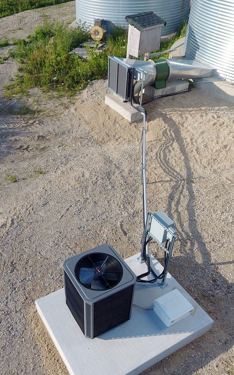 This photo shows a heat pump drying system installed on a grain bin. The components of the system are labeled, including the bin fan, the heat pump condenser coil in front of the fan, the heat pump compressor and evaporator module, and the refrigerant piping between the coil and the compressor.