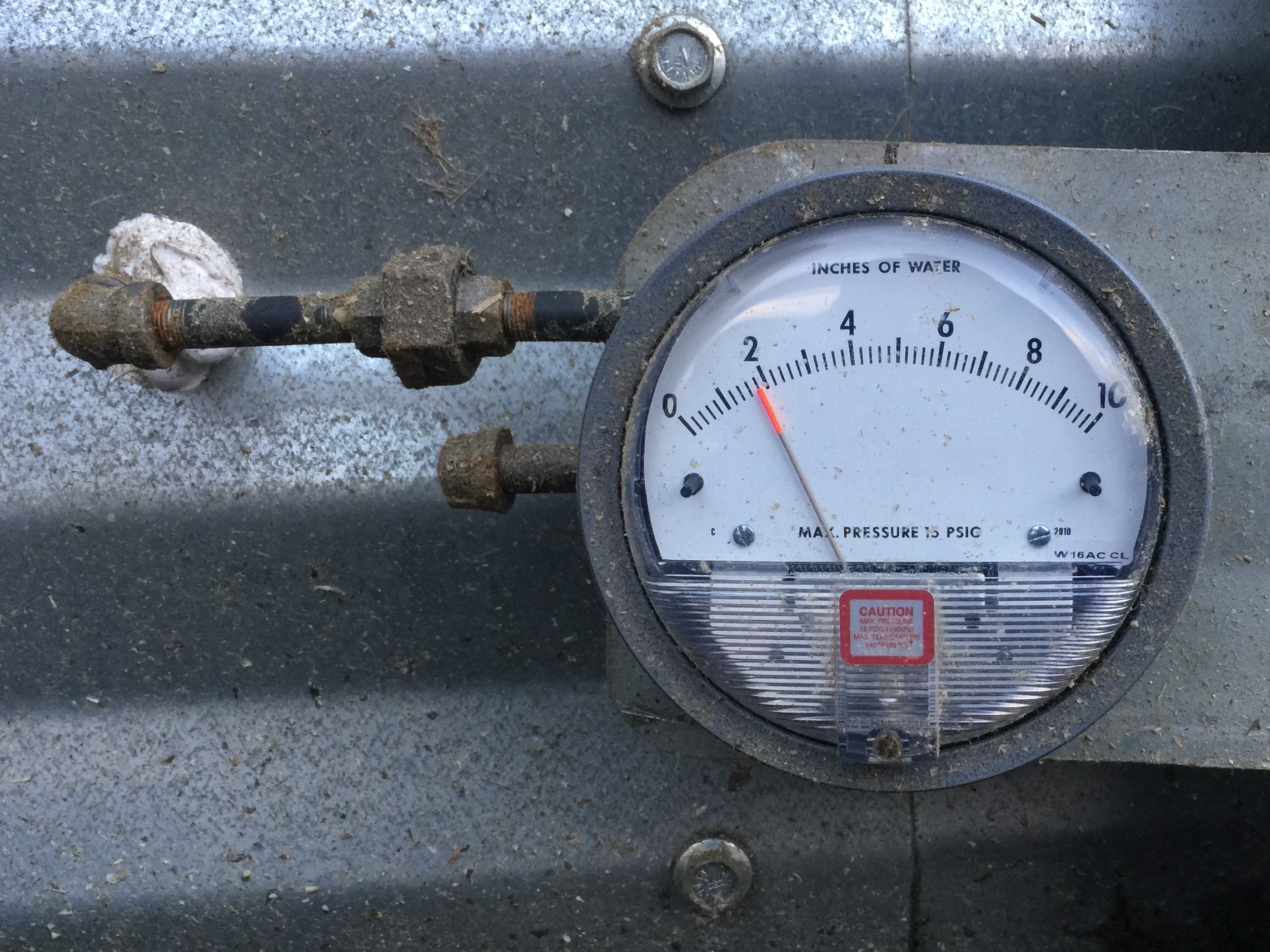 This photo shows a pressure gauge installed on a grain bin, to measure static pressure created by the fan. The gauge is round with a needle that measures pressure in inches of water.