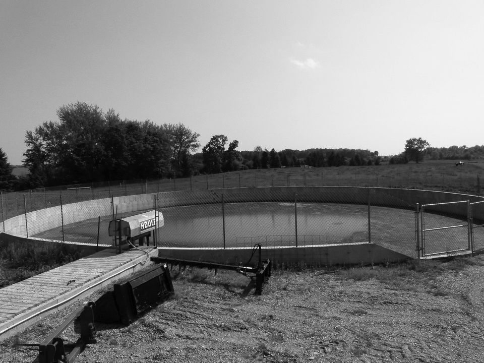Photo d’une structure d’entreposage de fumier circulaire enterrée, en béton. Le réservoir est entouré d’une clôture grillagée et situé à côté d’un champ de maïs.