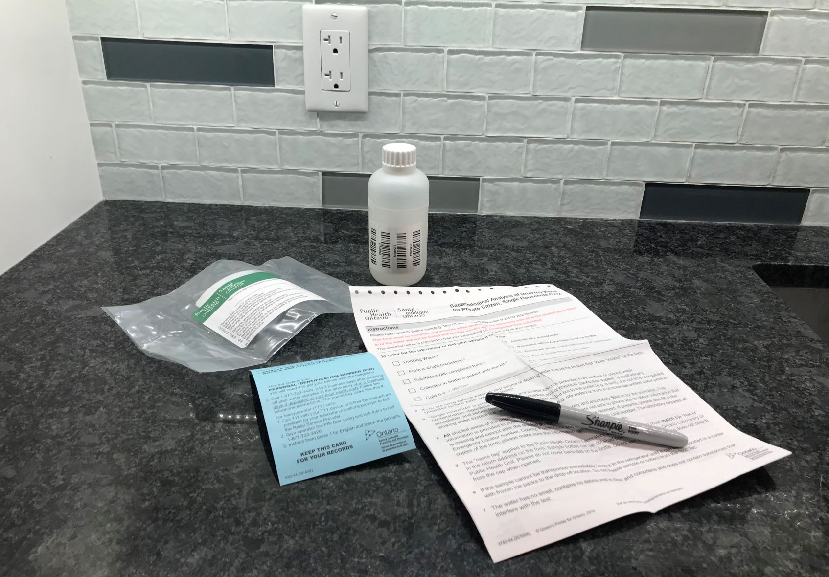 Une trousse d’analyse d’eau de puits privé sur un comptoir, avec une bouteille d’échantillonnage, un formulaire et un document donnant la marche à suivre et un stylo