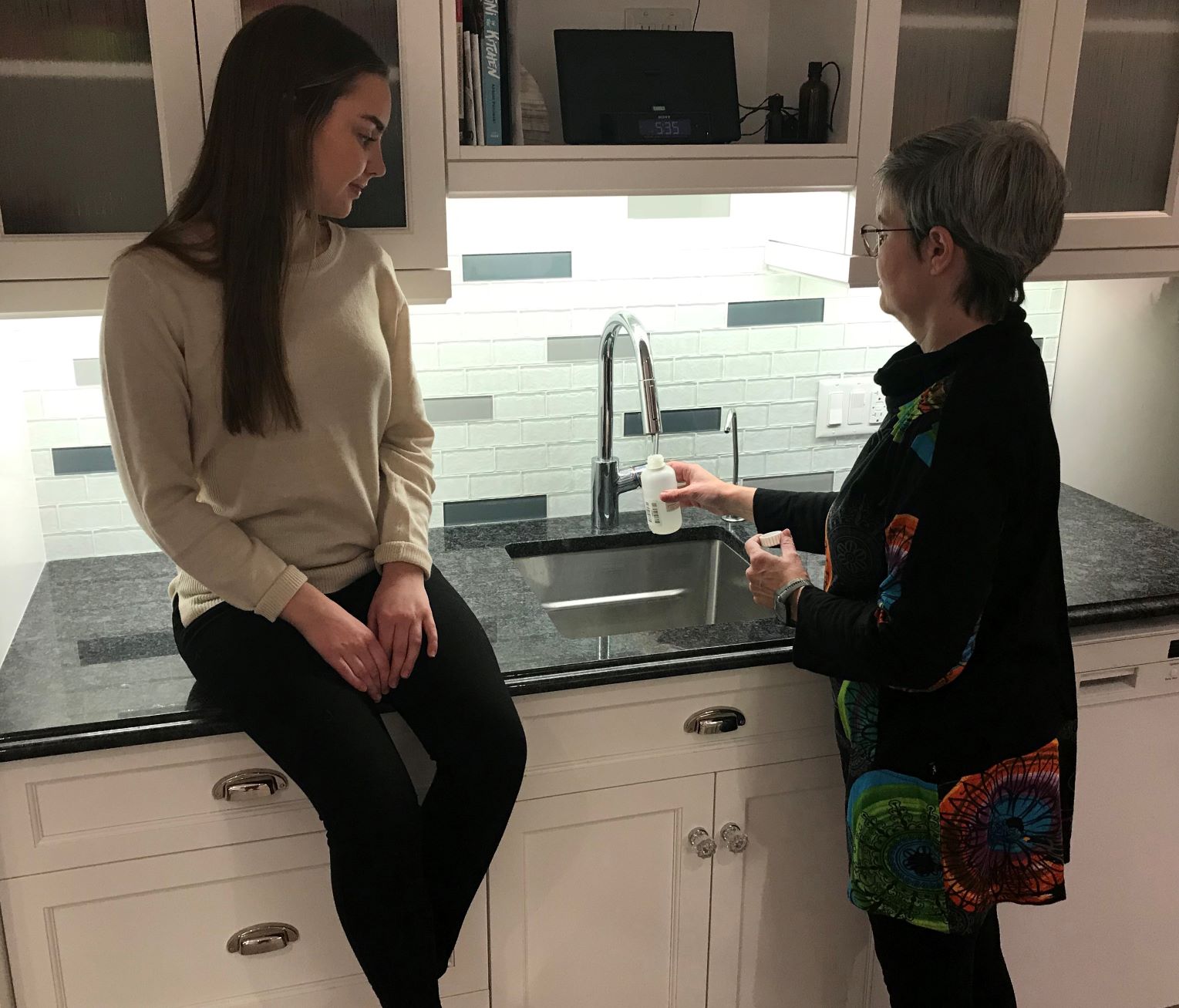 Une personne qui prélève un échantillon d’eau à partir d’un robinet de cuisine à l’aide de la bouteille fournie dans la trousse d’échantillonnage d’eau de puits. Une autre personne est assise sur le comptoir et regarde celle qui prend l’échantillon