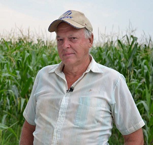 Male farmer wearing a baseball cap standing in front of a corn field.
