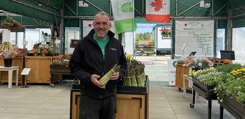Morris Gervais from Barrie Hill Farms holds asparagus while he stands in front of an asparagus display that is inside the farm store. There is a Canada flag and Foodland Ontario flag in the background. Fresh produce can be seen on the right side of the image and in the back left corner.