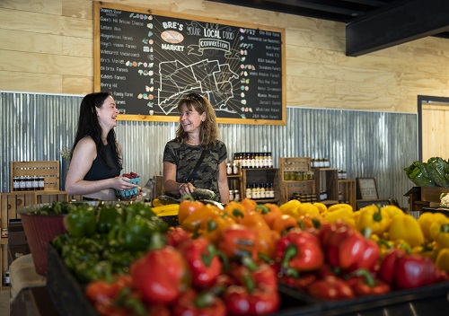 An image of two ladies standing in front of a map of local products offered at Bre’s Fresh Market in Tillsonburg. The ladies are standing in front of local fresh yellow and red peppers and leafy greens.