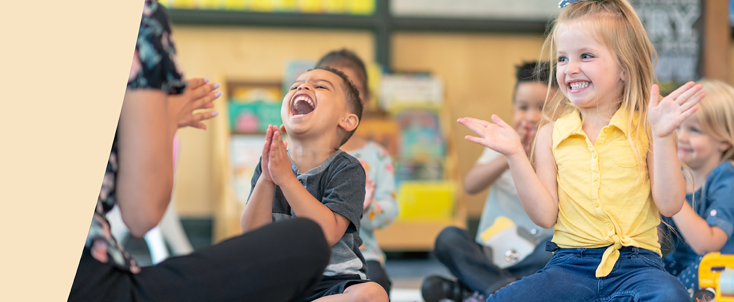 Young children clapping and smiling while sitting on a classroom carpet with an early childhood educator.
