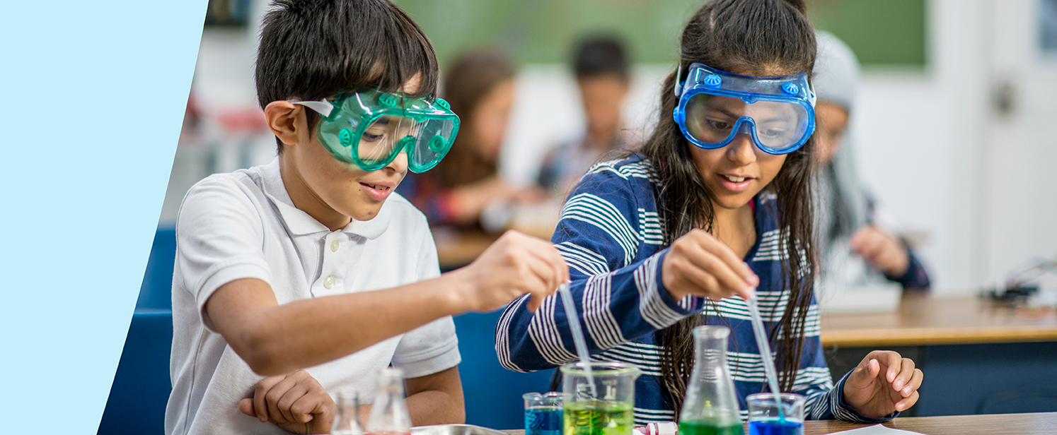 Students in a science classroom wearing safety goggles as they use pipettes and beakers filled with colourful liquids.