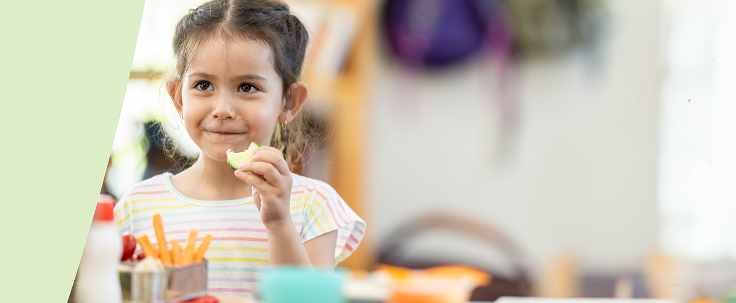 A young girl biting into a cucumber slice with her lunch in front of her.