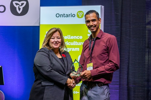 Minister of Agriculture, Food and Rural Affairs presents Anthony Chum with an Excellence in Agriculture award for the work that Moose Cree First Nations is doing with community gardens in Moose Factory Ontario. They are standing in front of a banner and shaking hands. This photo was taken at the awards presentation.