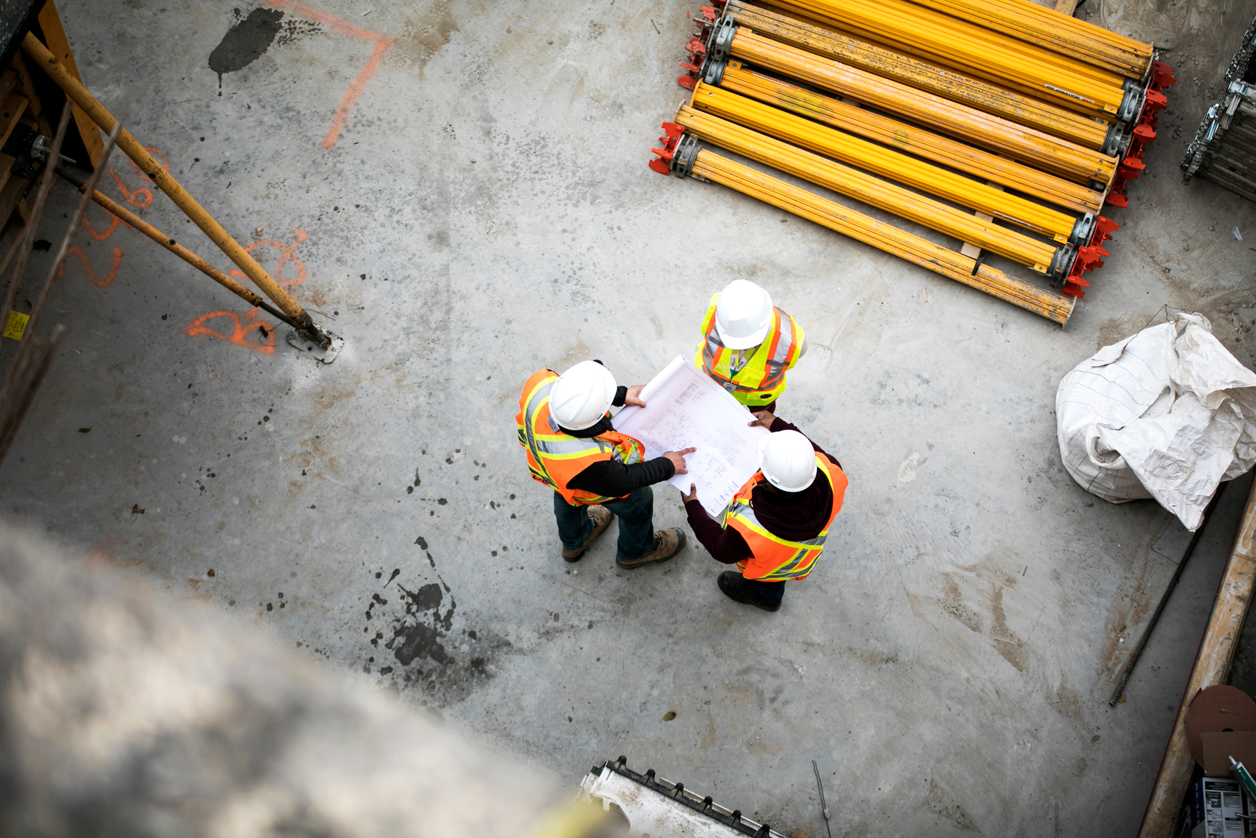 Vue aérienne de trois ouvriers du bâtiment en tenue de sécurité et casqués discutant de plans sur un chantier, avec des échafaudages jaunes à proximité. 