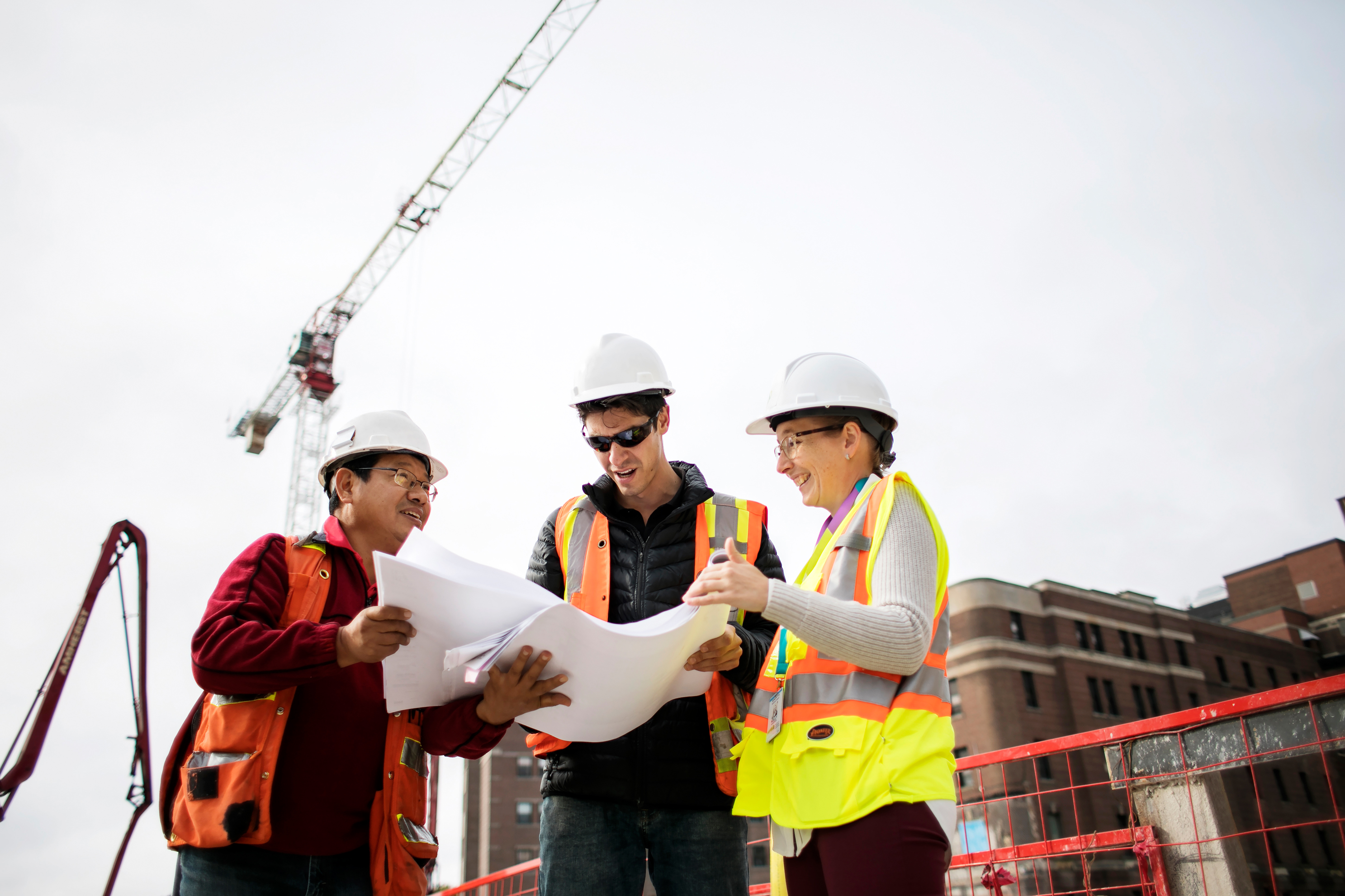 Three people at a construction site, wearing safety vests and hard hats, looking at a blueprint together