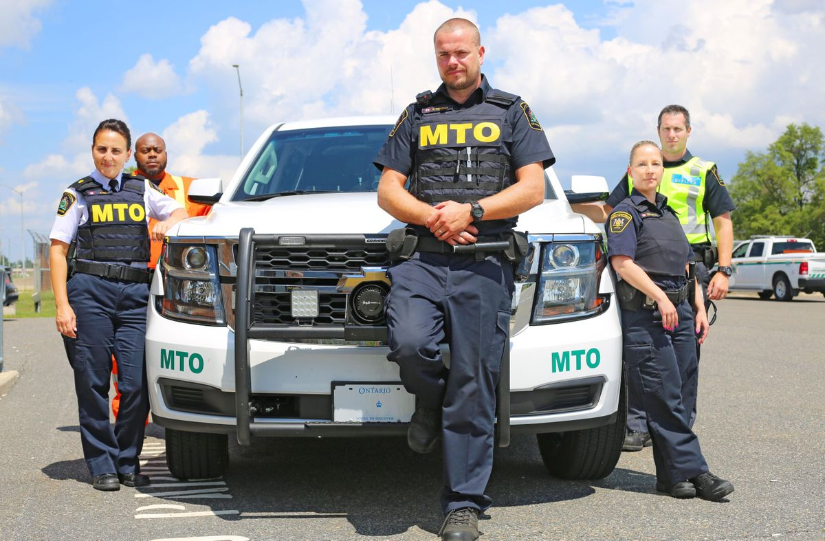 Five MTO transportation enforcement officers in uniform standing by a vehicle.