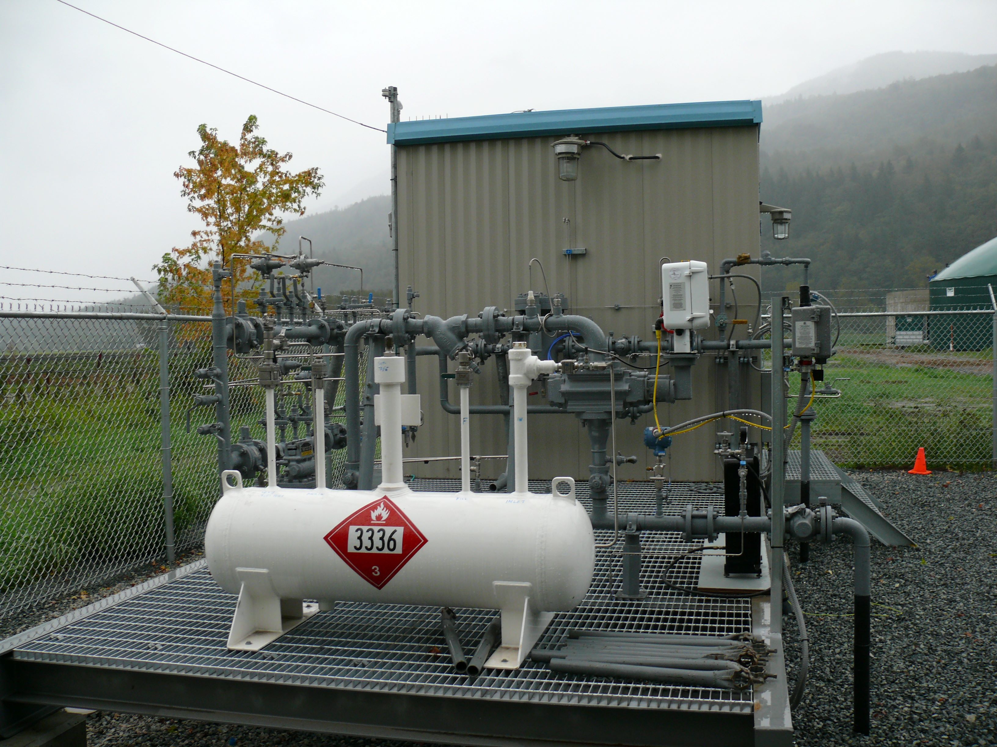a horizontal, white propane tank in front of a small building with natural gas pipelines in between