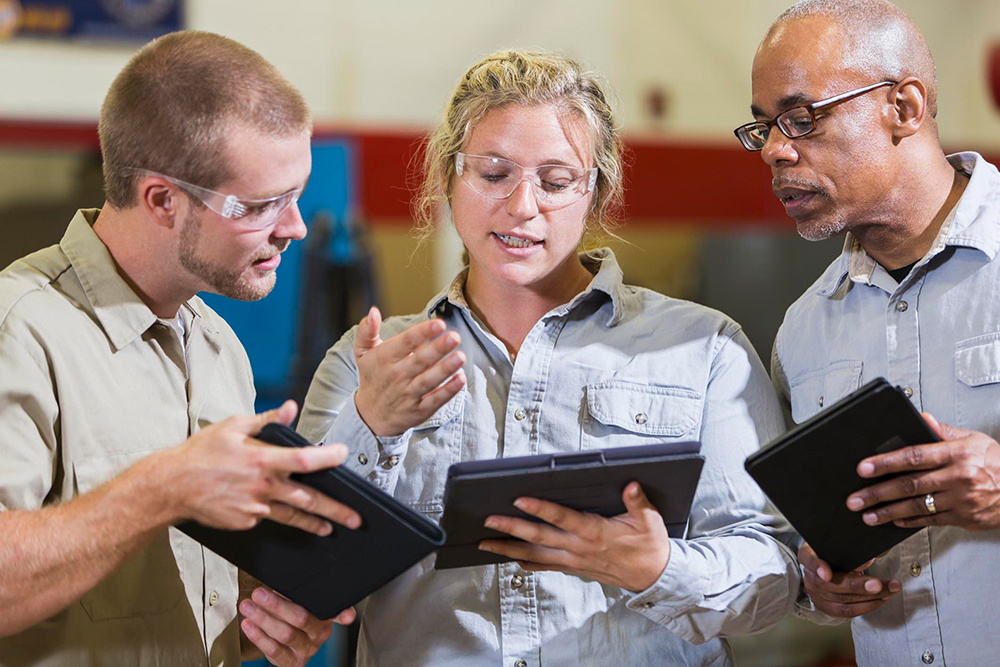 Three workers in a manufacturing environment are standing close together, each holding a tablet. They are engaged in a discussion about what they are seeing on their tablets.