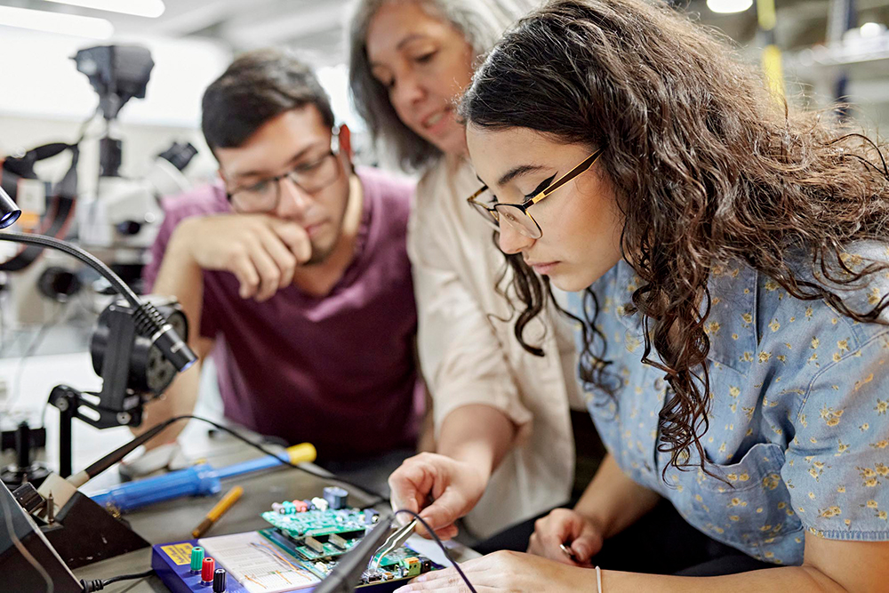 Two students with their teacher are working together on an electronics project. They are gathered around a table with various electronic components and a circuit board. The teacher is pointing to a part of the circuit board.