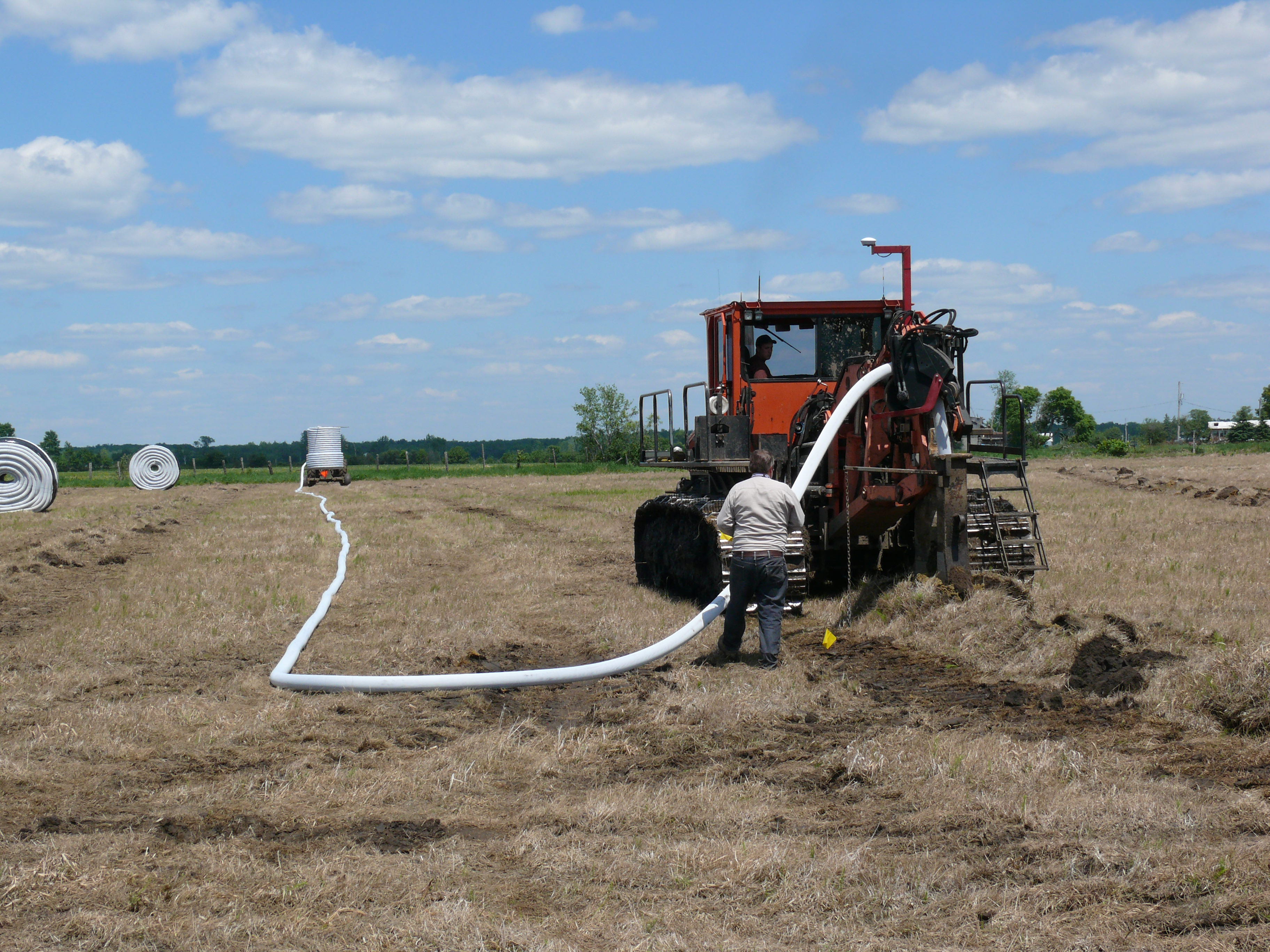 A drainage tile machine installing white drainage tile in a field, with a man standing behind the machine.