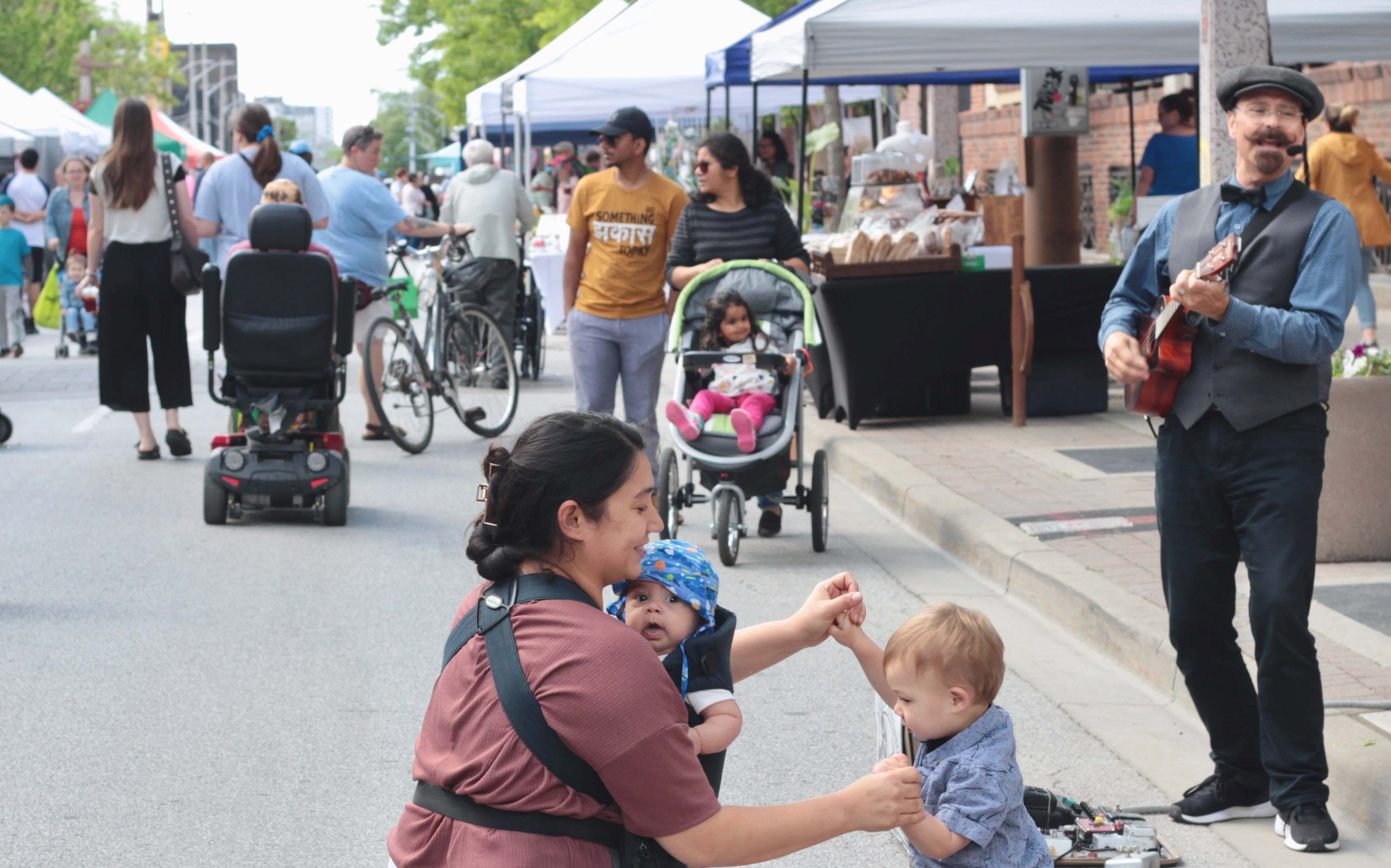 An image from the Downtown Windsor Farmers’ Market with a mom and two young kids front and centre. A busker playing guitar