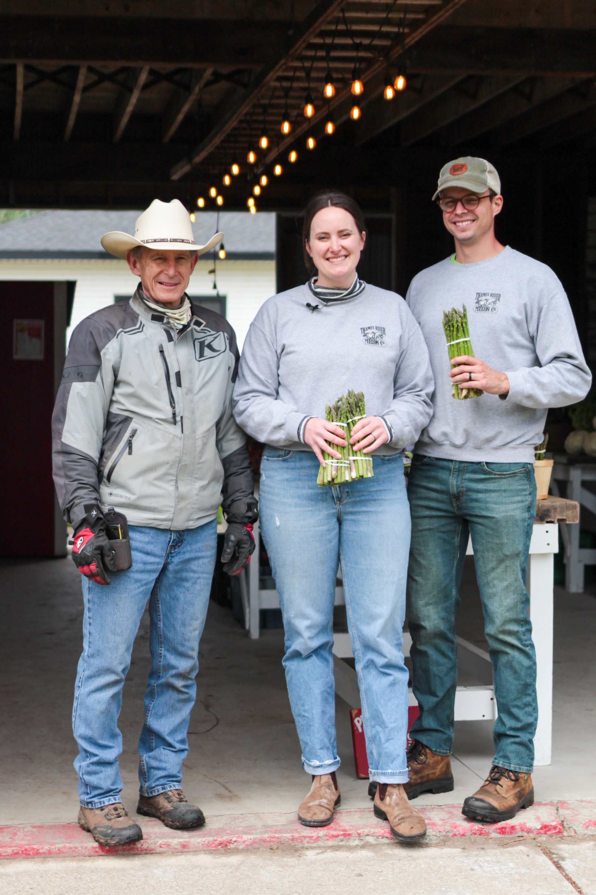 An image of 3 people standing together. The middle person and person on the far right are holding asparagus.