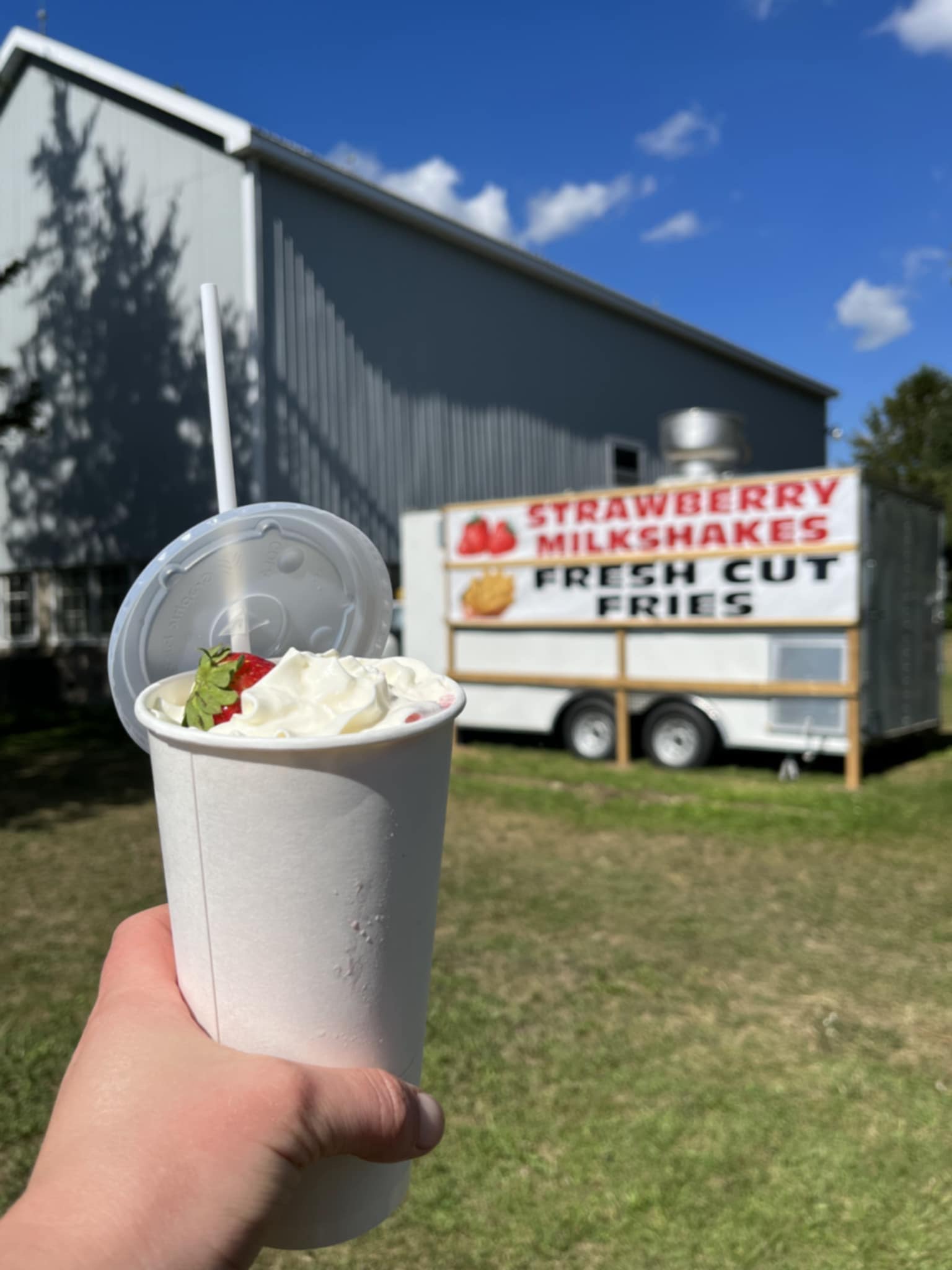 An image of hand holding a milkshake in the foreground. In the background there is a white food truck.