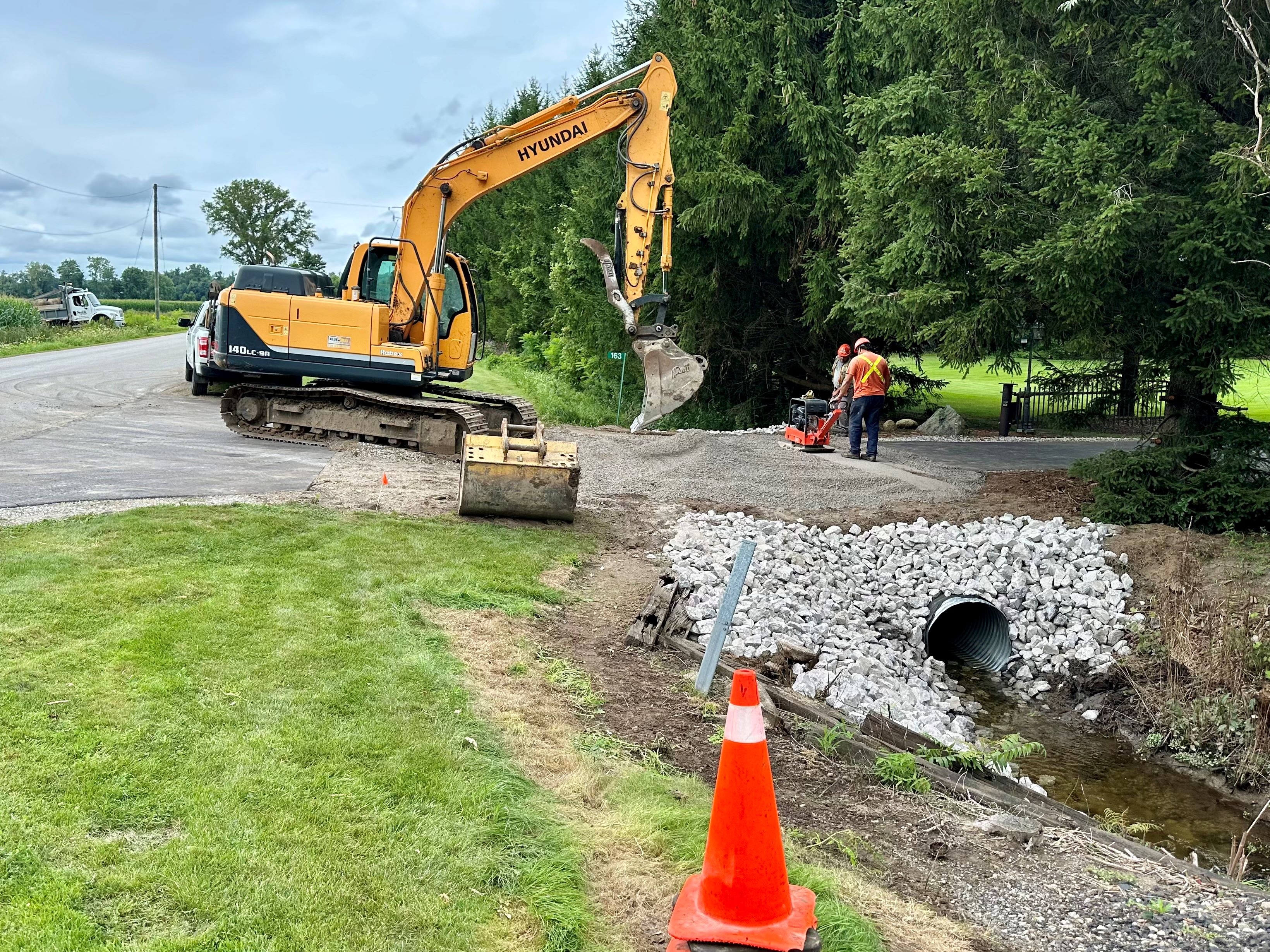 An excavator working on a drain that runs under a driveway.  There are workmen to the right of the drain.