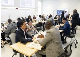 Photo of a large room where several groups of people are seated around tables, engaging in pair or small‑group discussions; in the background, a presentation screen displaying content is visible
