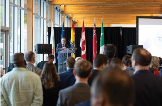 Photograph from the OIF trade mission co‑sponsored by Ontario: Dominic Mailloux, President of Fédération des gens d’affaires de l’Ontario, standing at a lectern addressing the audience, with several provincial and national flags behind him and a projection screen partially visible to the right.”