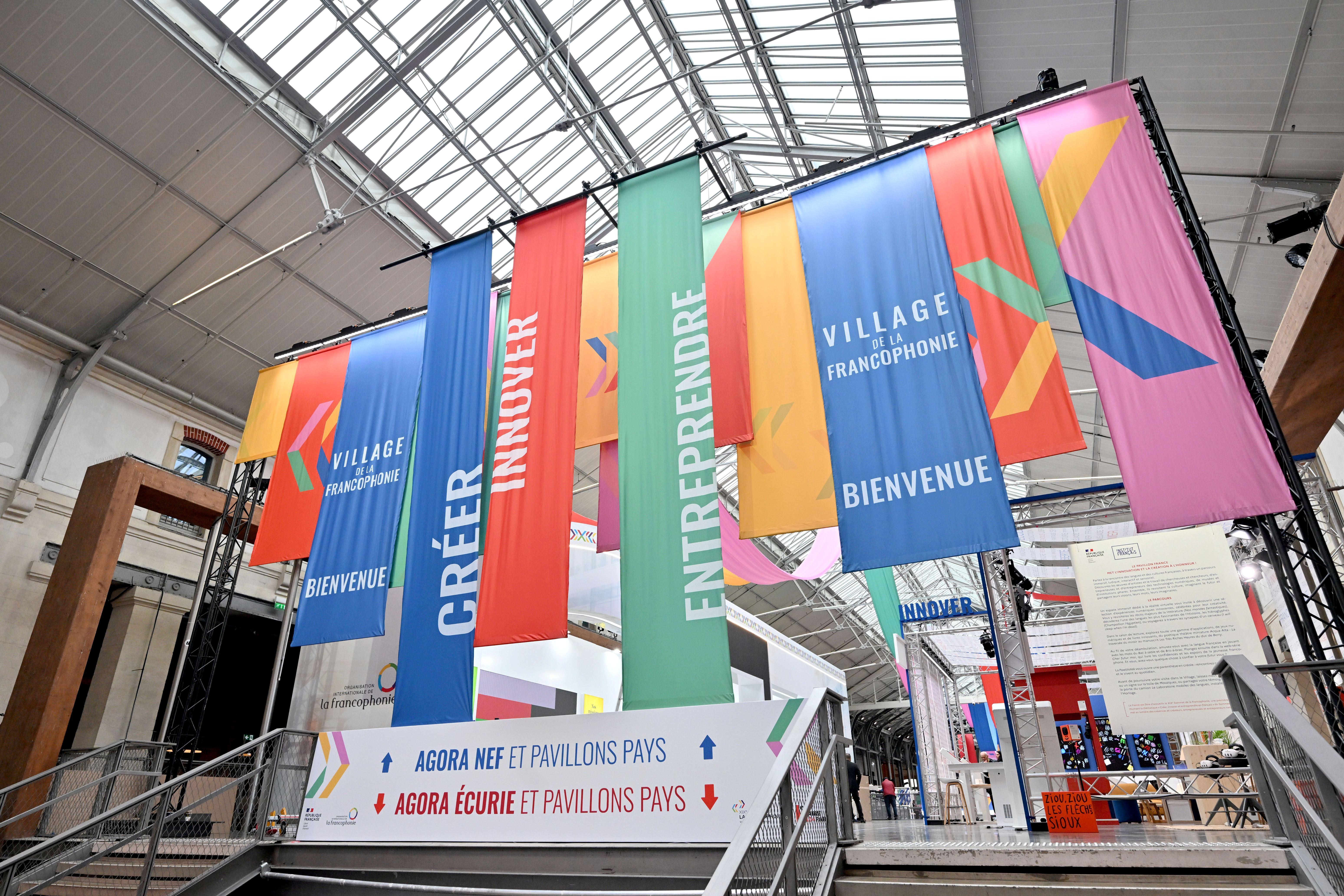 Photograph of the Village de la Francophonie exhibition space: an indoor area with several colorful vertical banners hung from the ceiling reading ‘CRÉER’ (blue), ‘INNOVER’ (red), ‘ENTREPRENDRE’ (green), and ‘VILLAGE DE LA FRANCOPHONIE – BIENVENUE’ (blue), with directional signs below pointing to various pavilions