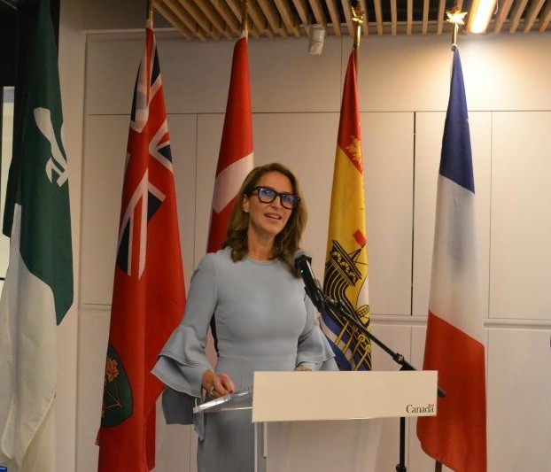 Photograph of Minister Caroline Mulroney wearing a light blue dress and glasses, standing behind a white podium labeled ‘Canada’ and addressing an audience; in the background, five flags are displayed: Franco‑Ontarian, Ontario, Canada, New Brunswick, and France