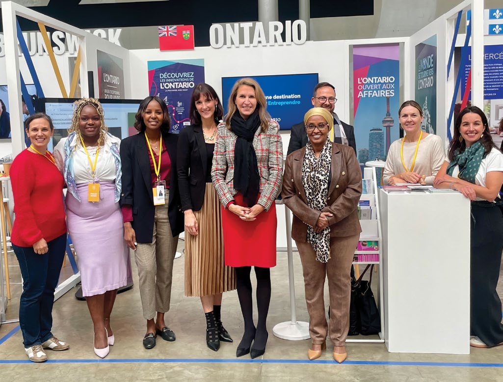 Photograph of members of the Ontario delegation standing in a row in front of the Ontario booth at the Village de la Francophonie; at the center are Minister Caroline Mulroney and Deputy Minister Roda Muse, inside a large exhibition hall with ‘Ontario’ and ‘Discover Ontario’ signage in the background.”