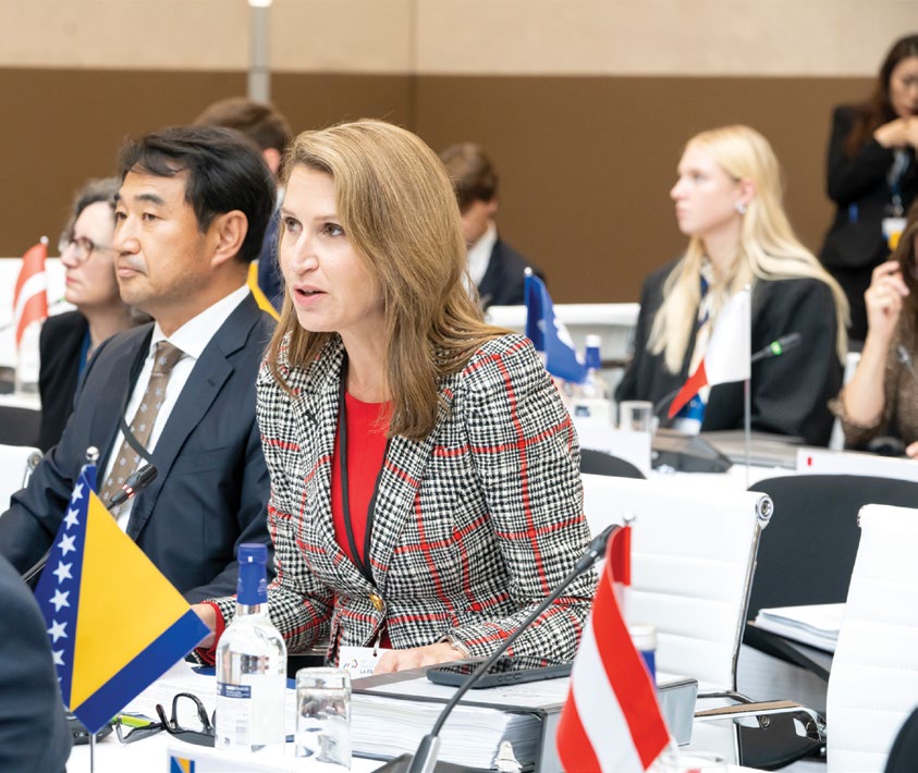 Photograph of Ontario’s Minister of Francophone Affairs, Caroline Mulroney, speaking as part of the official program of the Francophonie Summit: she is seated at a conference table alongside other international delegates, with several national flags visible in the foreground and background