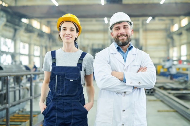 Two people at a factory wearing hard hats and overalls.