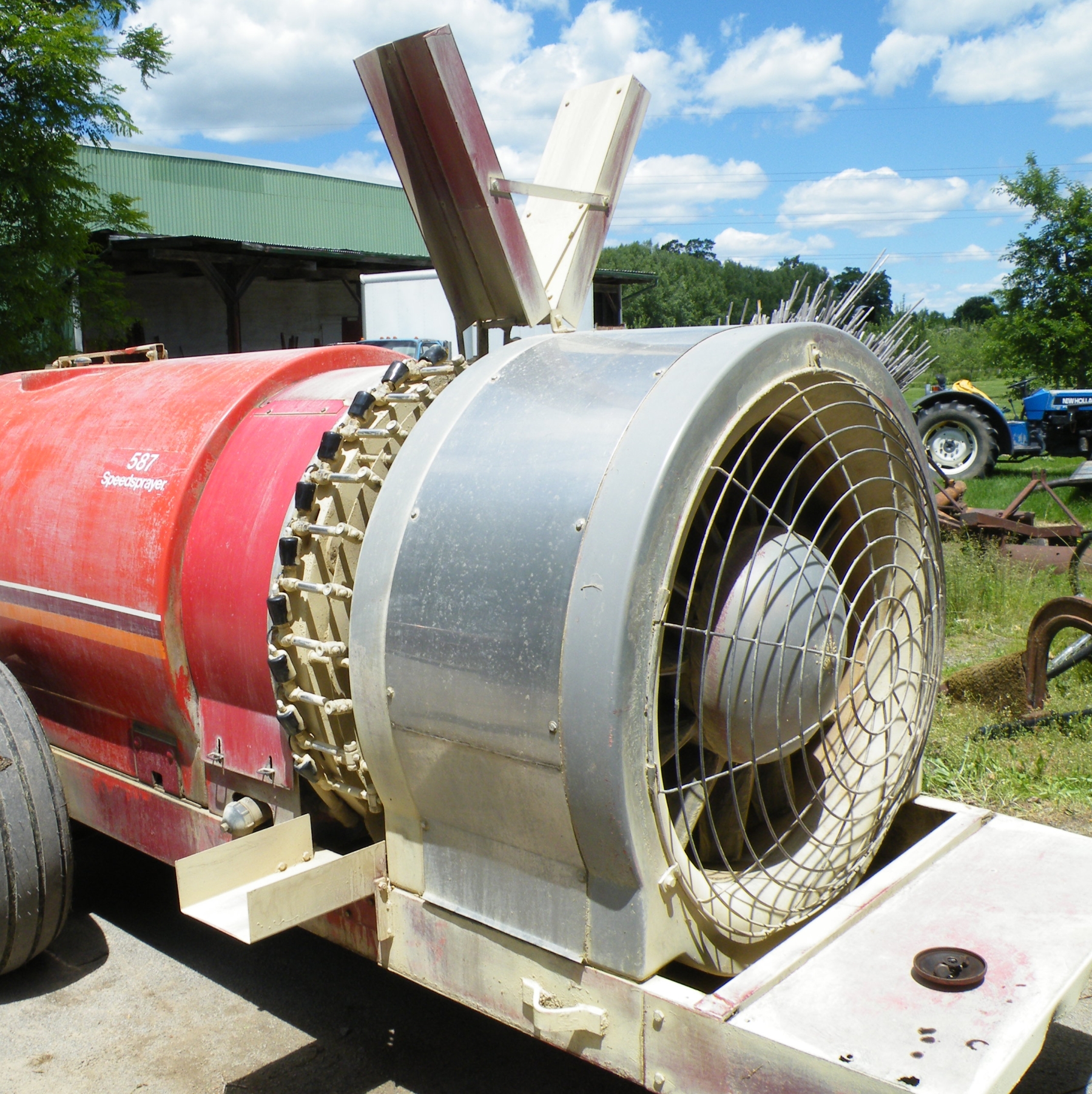 Red airblast sprayer, with a large metal fan and nozzles. There are metal deflectors in a V shape that deflect the spray coming from the nozzles that are located before the metal fan.