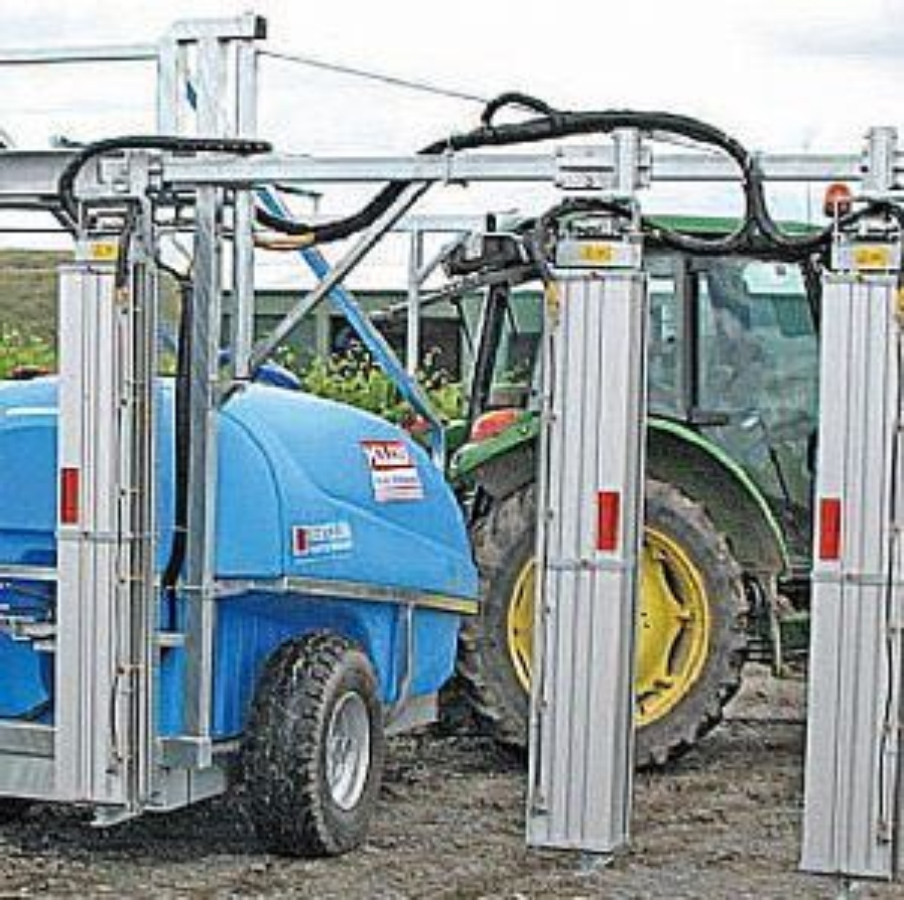 A tangential sprayer. Features three metal columns with tubes attached to a blue sprayer being pulled behind a tractor.