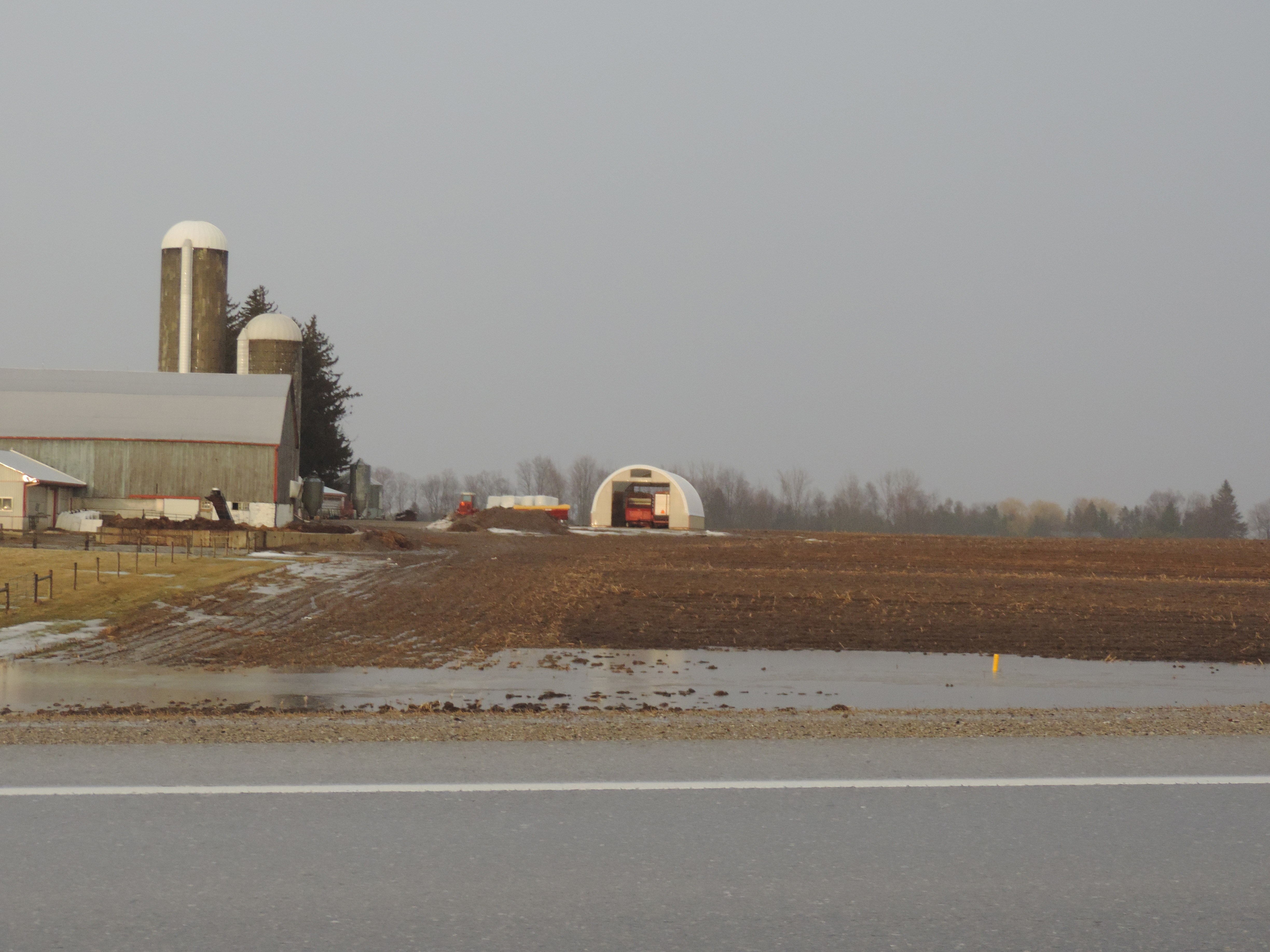 une étable à côté d’un champ dénudé recouvert d’un peu de neige en train de fondre. On voit aussi une zone où l’eau s’est accumulée autour d’une entrée de drain qui longe une route à l’avant-plan