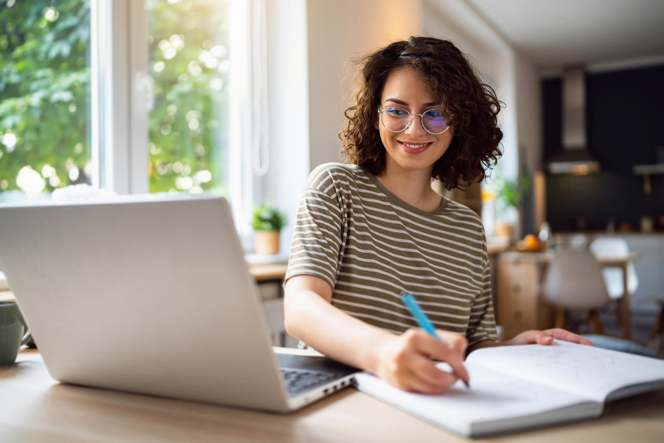 Une personne assise à un bureau dans un bureau à domicile qui écrit dans un cahier et travaille sur un ordinateur portable.